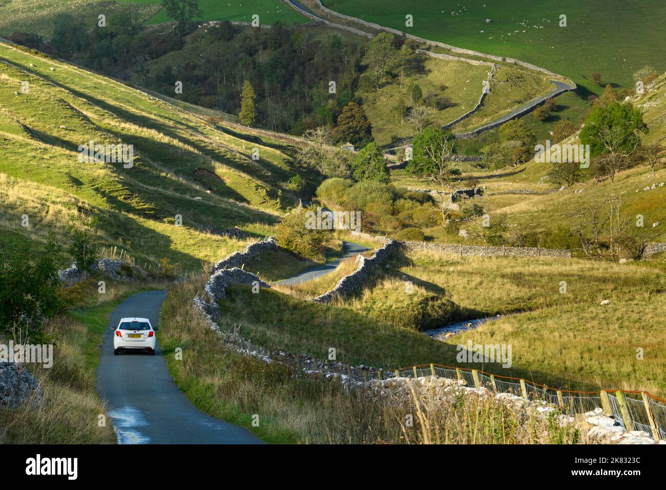 1 coche en un tranquilo y ondulante carril campestre (paisaje montañoso iluminado por el sol, empinado ascenso, arroyo) - cerca de Kettlewell, Yorkshire Dales, Inglaterra, Reino Unido. Foto de stock