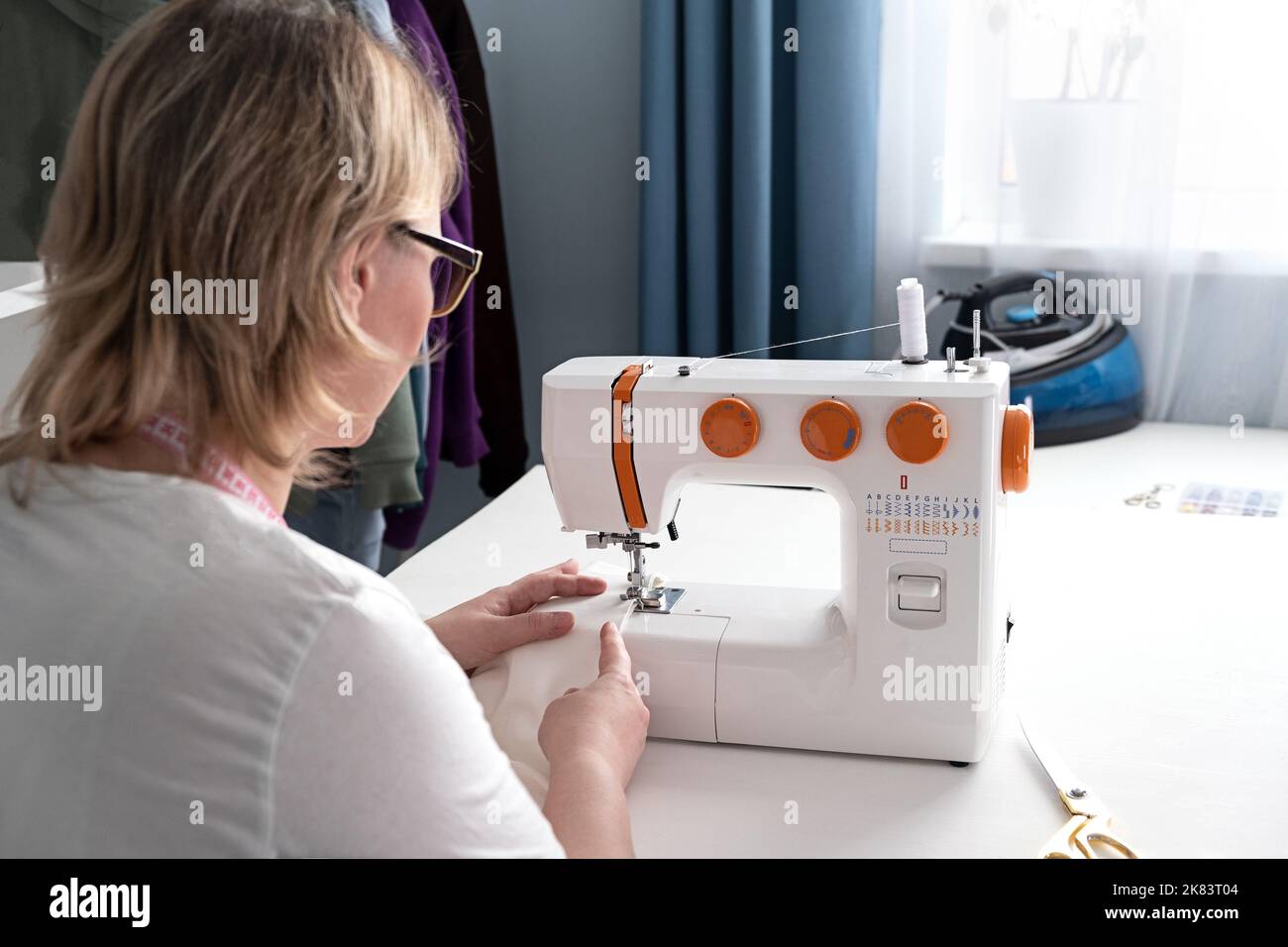Mujer de mediana edad real sastre la ropa con una máquina de coser en casa.  Trabajo en costureras. Hacer ropa a mano en el interior de la casa. La  mujer se sienta