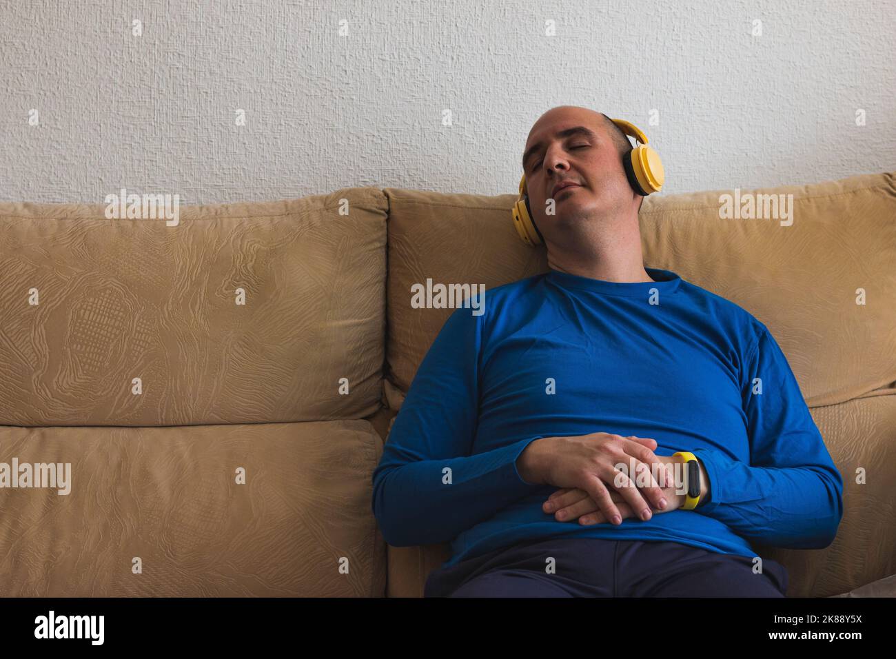 Un hombre caucásico calvo vestido con una camiseta azul de manga larga está dormido sentado en un sofá mientras lleva auriculares amarillos. Foto de stock