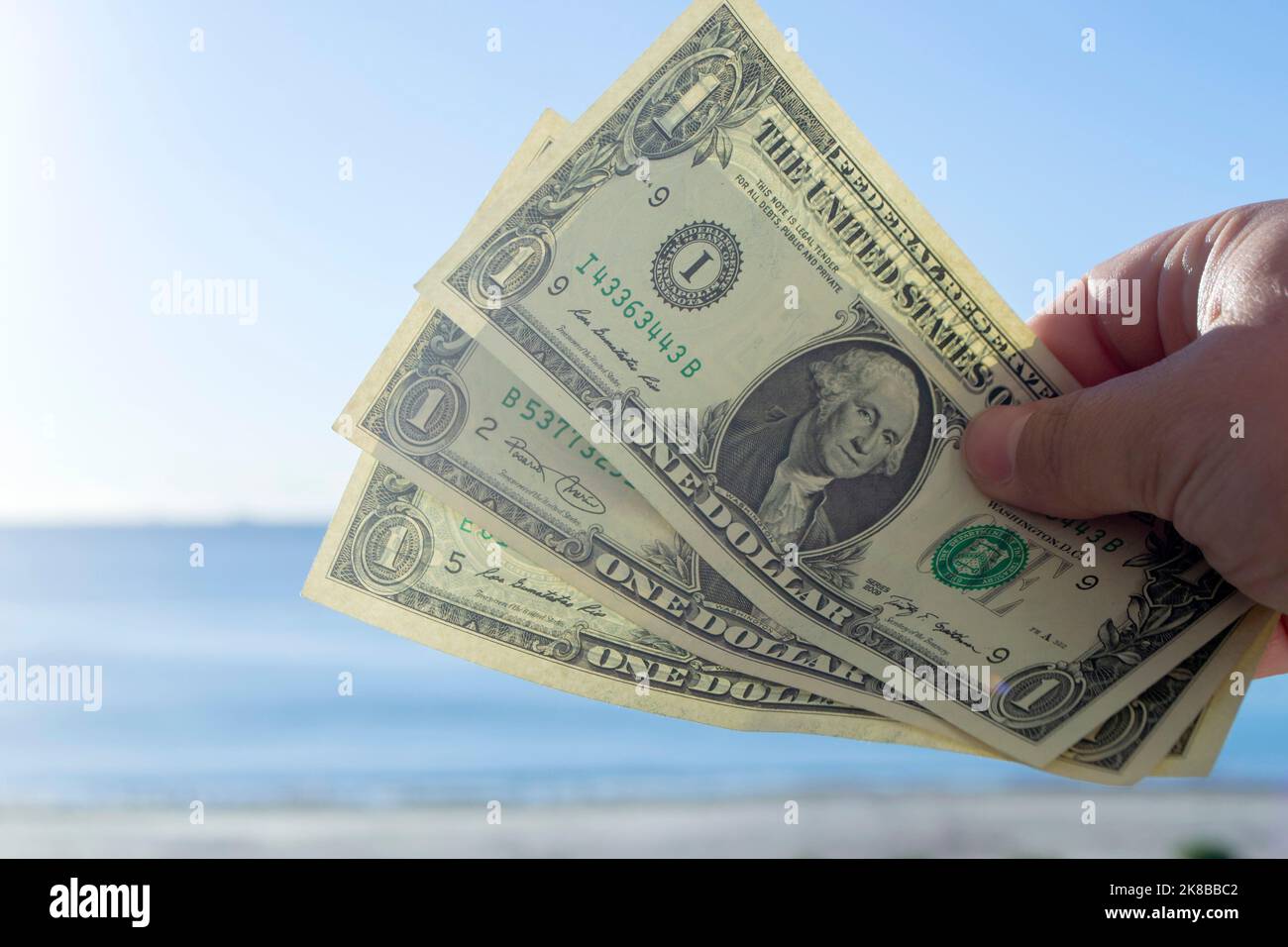 Un hombre sosteniendo tres billetes de un dólar en su mano contra el fondo del mar, la playa de arena y el cielo azul en el soleado día de verano. El concepto de dinero, finanzas, dinero, viajes, turismo, vacaciones, descanso Foto de stock