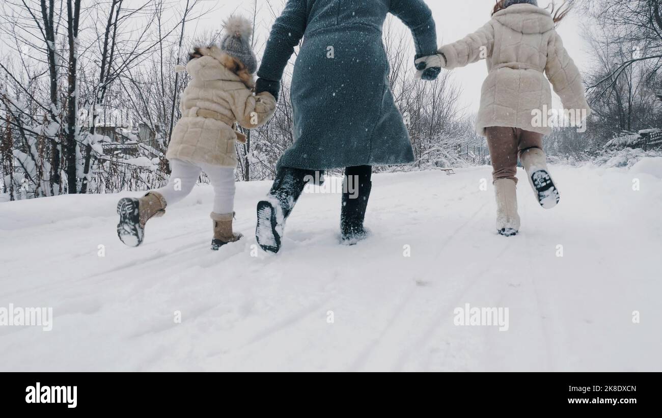 actividad familiar de invierno al aire libre. vista posterior. Una mujer feliz con 2 hijas corre en un camino nevado, en el bosque, durante la nevada. La familia se divierte, pasando tiempo juntos en un día nevado de invierno. Cámara lenta. Fotografía de alta calidad Foto de stock