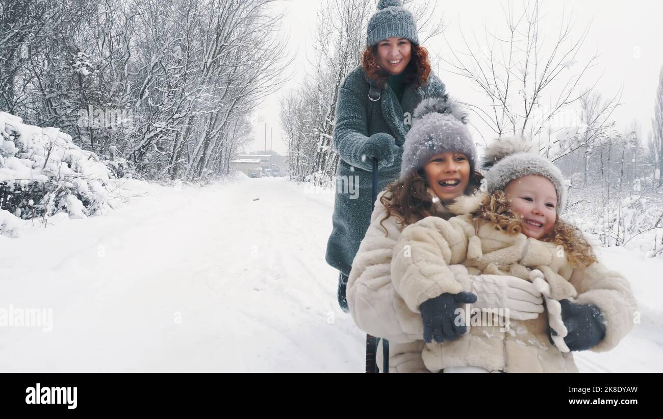 paseos en trineo familiares en invierno. actividades al aire libre en invierno. Feliz, familia riendo, una mujer con 2 hijas están disfrutando de trineos en la carretera nevada, en el bosque, durante la nevada. La familia se está divirtiendo, pasando tiempo juntos el día nevado del invierno. Cámara lenta. Fotografía de alta calidad Foto de stock