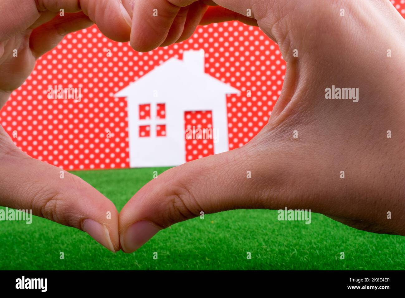 Casa de papel en el centro de una mano que forma un corazón sobre un fondo rojo de puntos blancos con hierba Foto de stock