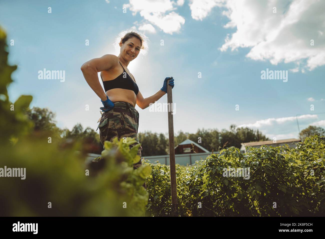 Mujer madura sonriente sosteniendo pala de pie con la mano en la cadera en el jardín de verduras Foto de stock