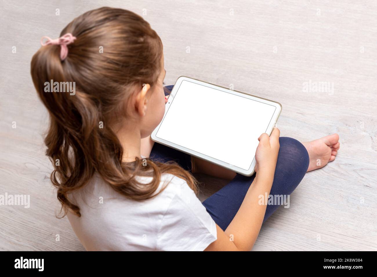 Niña sentada en el suelo y sosteniendo una tableta blanca con pantalla aislada para promocionar videojuegos, sitios web o aplicaciones Foto de stock