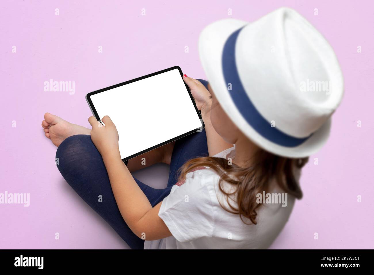 La niña con un sombrero blanco está sentada en un suelo rosa y sostiene una tableta con pantalla aislada. Maqueta de la tableta Foto de stock