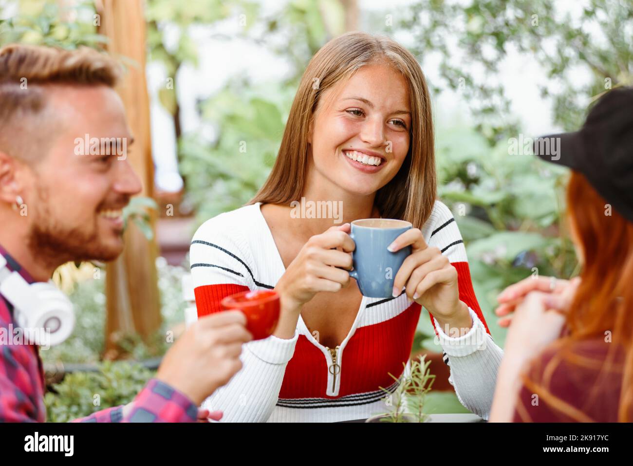 Mujer joven feliz sonriendo y escuchando a un amigo mientras beben café juntos el día de verano en el jardín Foto de stock