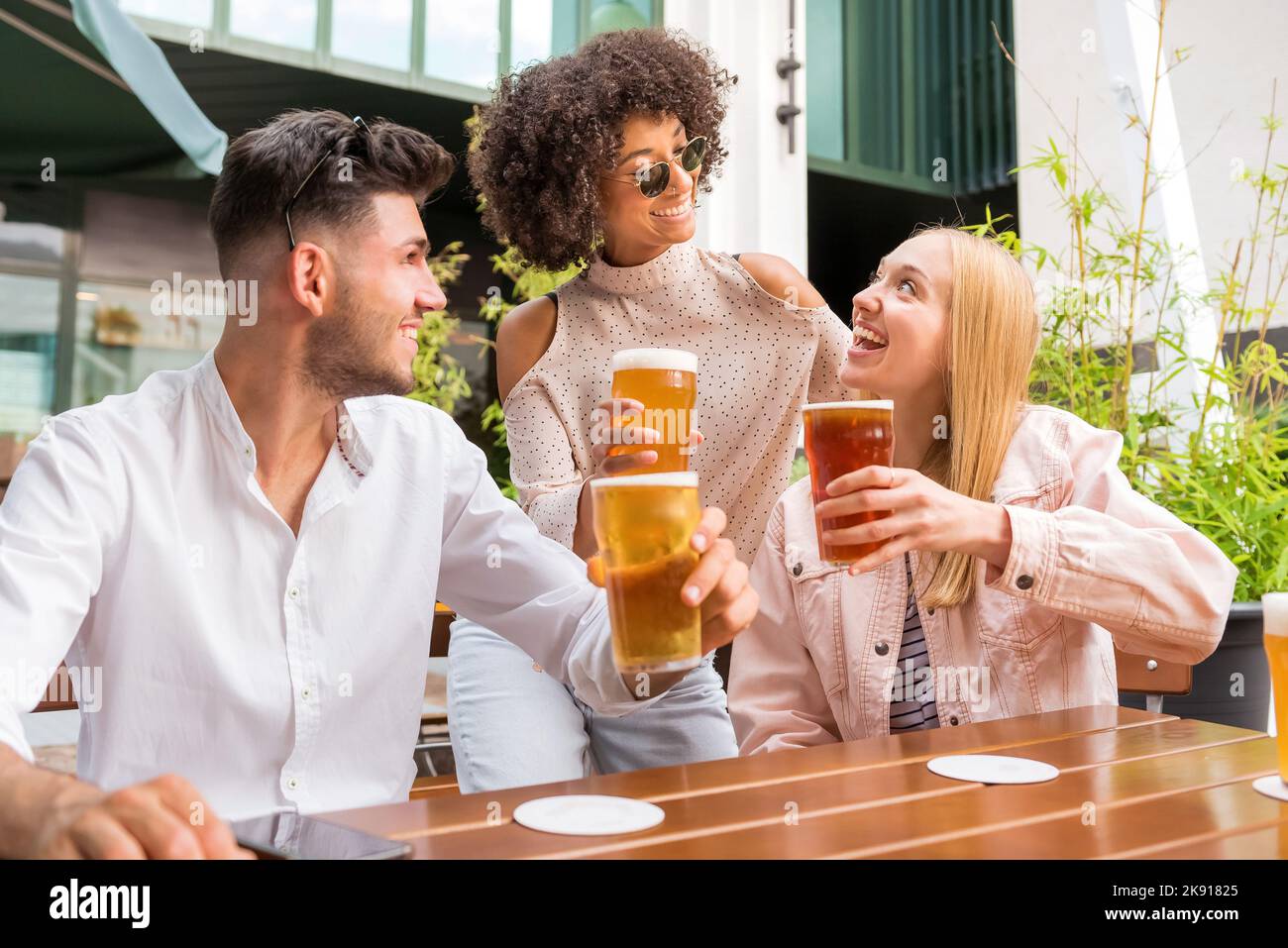 Amigos multirraciales positivos mirándose unos a otros y acercándose vasos de cerveza mientras se sientan juntos en una mesa en un pub al aire libre Foto de stock