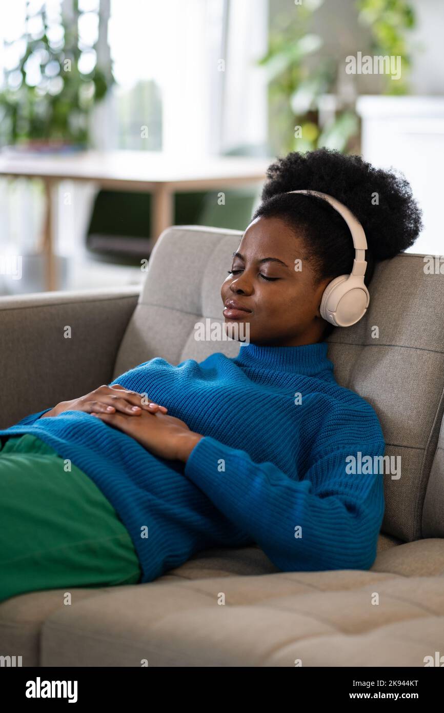 Una mujer afroamericana que descansa con unos auriculares con una leve sonrisa se encuentra en el sofá y se duerme con la música Foto de stock