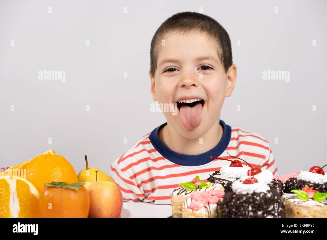 niño lindo de 5 años se sienta frente a frutas y pasteles y elige qué comer Foto de stock