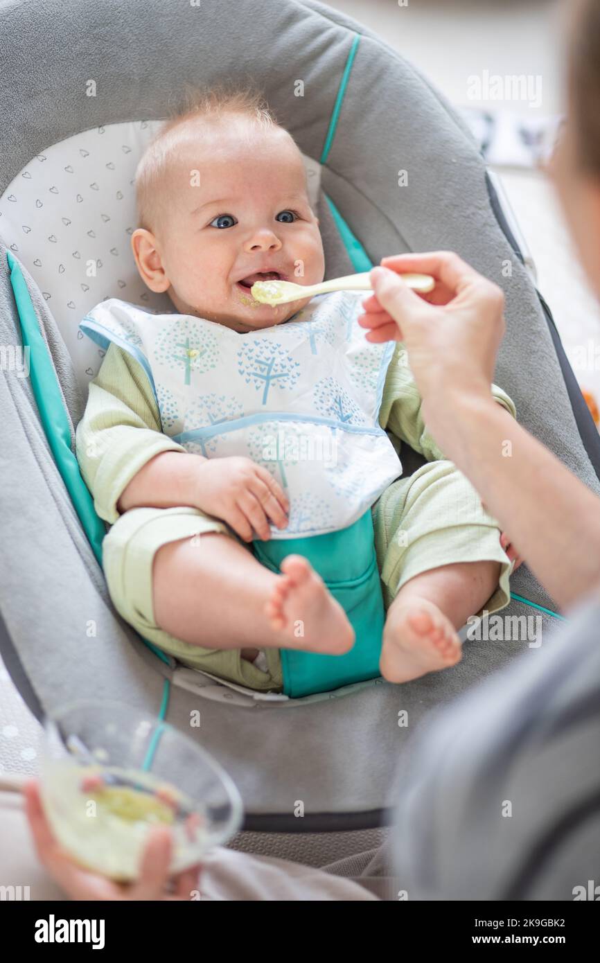 Cuchara madre alimentando a su bebé niño en silla de bebé con puré de frutas. Concepto de introducción de alimentos sólidos para bebés. Foto de stock