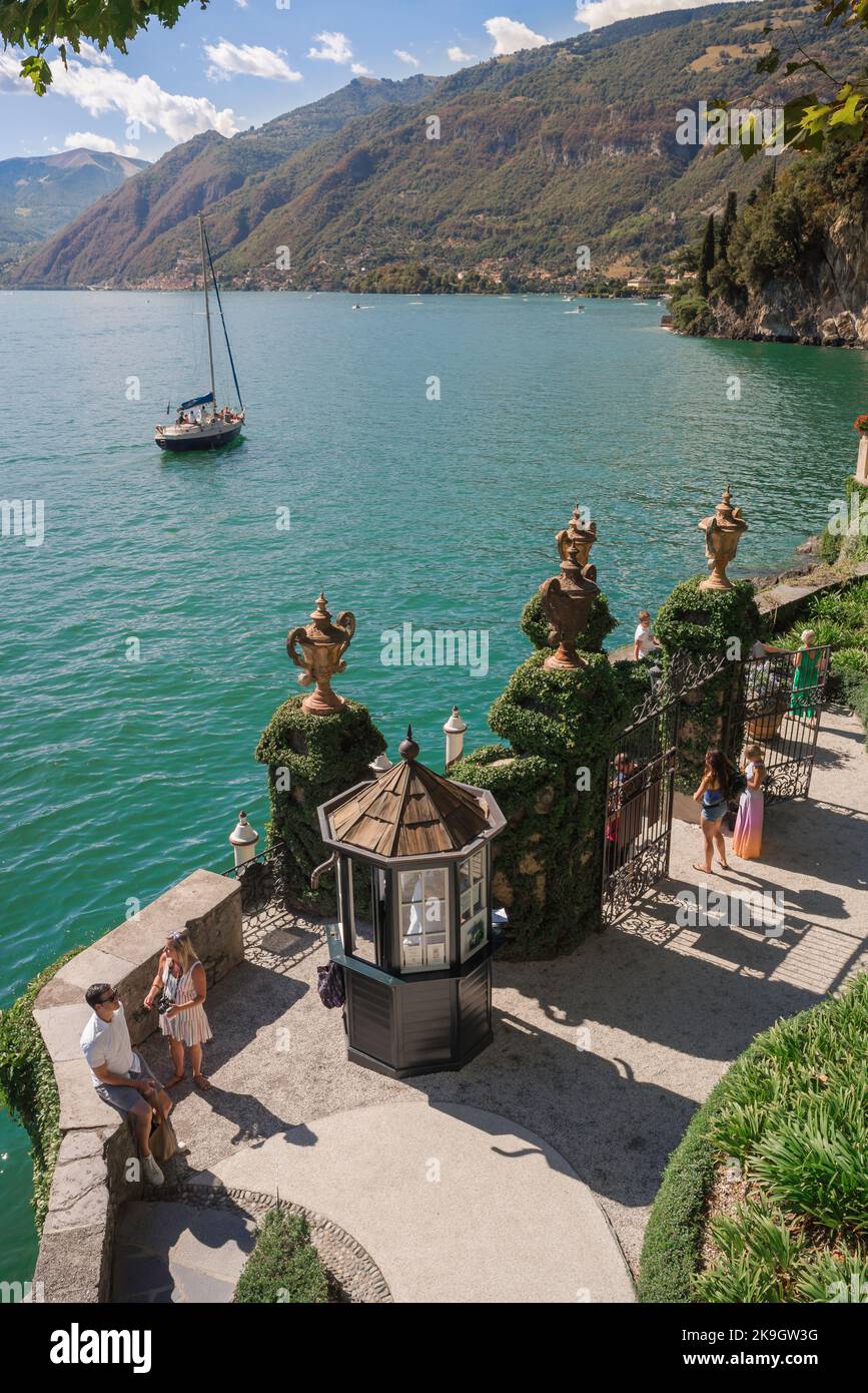 Italia gente del lago, vista en verano de la gente de pie en la terraza del lago de la Villa del Balbienello con vistas a un yate de vela Lago Como, Italia Foto de stock