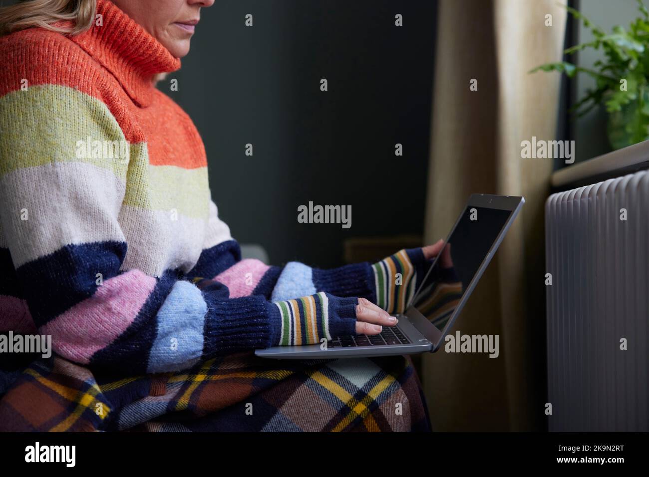 Mujer en guantes con computadora portátil tratando de mantenerse caliente por el radiador durante la crisis del costo de vida de la energía Foto de stock