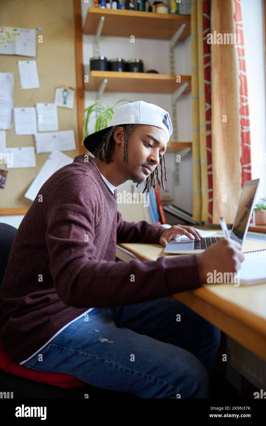 Estudiante universitario o universitario masculino con gorra de béisbol estudiando con computadora portátil en el escritorio de la habitación Foto de stock