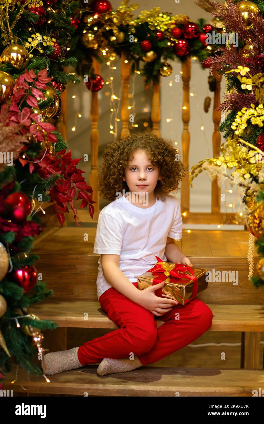 Bonita chica rizada en pijama sentada sobre escaleras de madera en estilo  loft de sala de estar. Escaleras decoradas para Navidad. El niño sostiene  una caja de regalo. Feliz Navidad an Fotografía