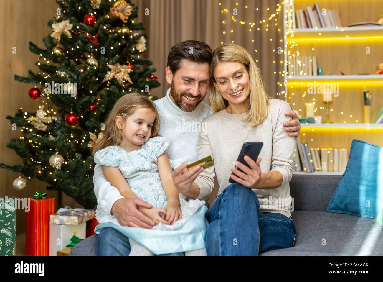 Feliz familia para las compras de Navidad en línea eligiendo regalos en la tienda en línea, el hombre y la mujer con el niño sentado en casa en el sofá cerca del árbol de Navidad que sostiene la tarjeta de crédito del banco. Foto de stock