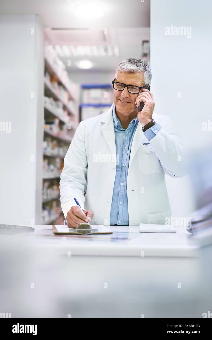 Buen día, cómo puedo ayudar. Un farmacéutico masculino maduro alegre que hace notas mientras que está en el teléfono en la farmacia. Foto de stock