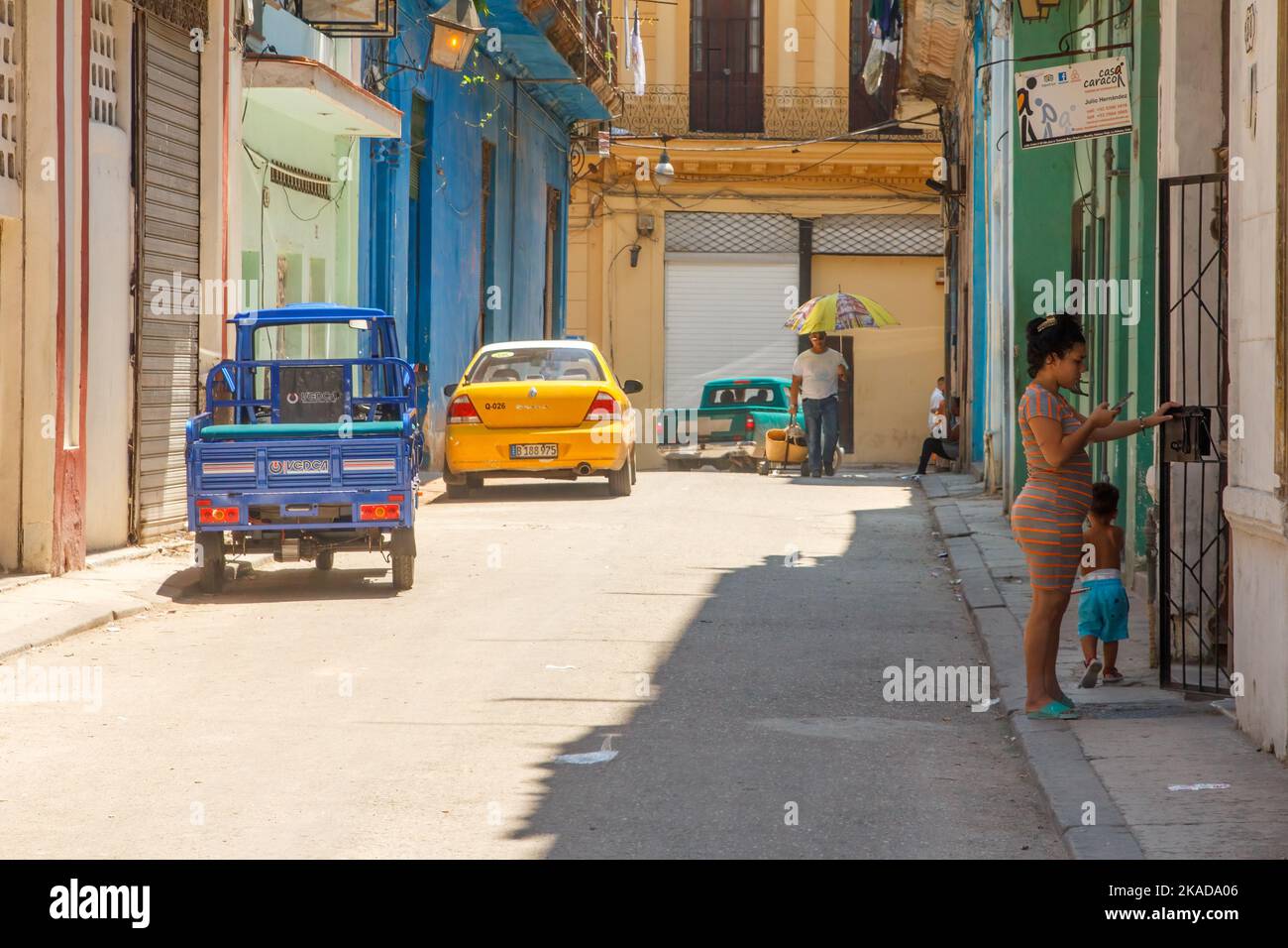 Una mujer cubana interactúa a la puerta de un pequeño negocio privado. Los vehículos modernos están estacionados en la calle de la ciudad. Foto de stock