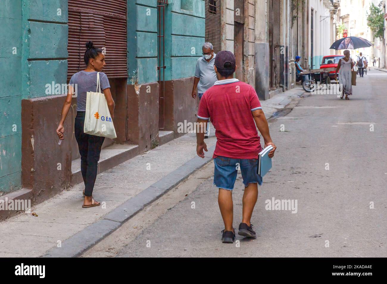 El estilo de vida de los cubanos mientras caminan por la calle de una ciudad. Un hombre que lleva libros habla con una mujer en la acera Foto de stock