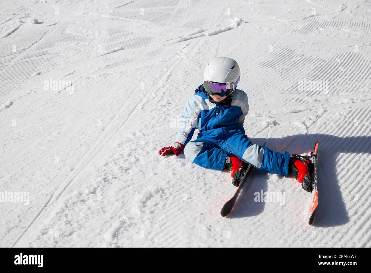 un niño cansado en un casco, gafas de esquí, esquís y monos de invierno se sienta sobre la nieve. Cayó en la nieve, se detuvo para descansar. Diversión invernal. Aprendizaje de niño pequeño Foto de stock