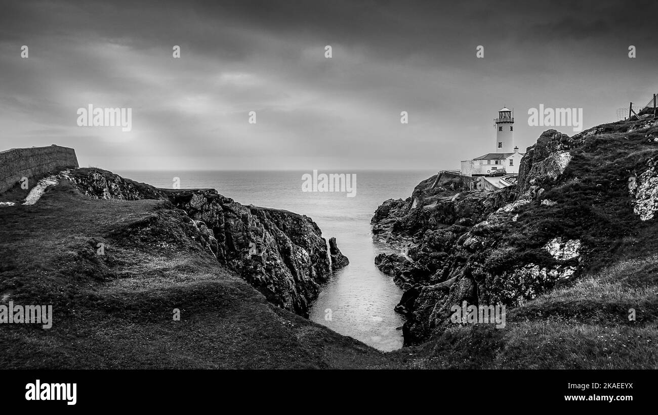 Un paisaje blanco y negro de mar, rocas y un faro en Irlanda. Foto de stock