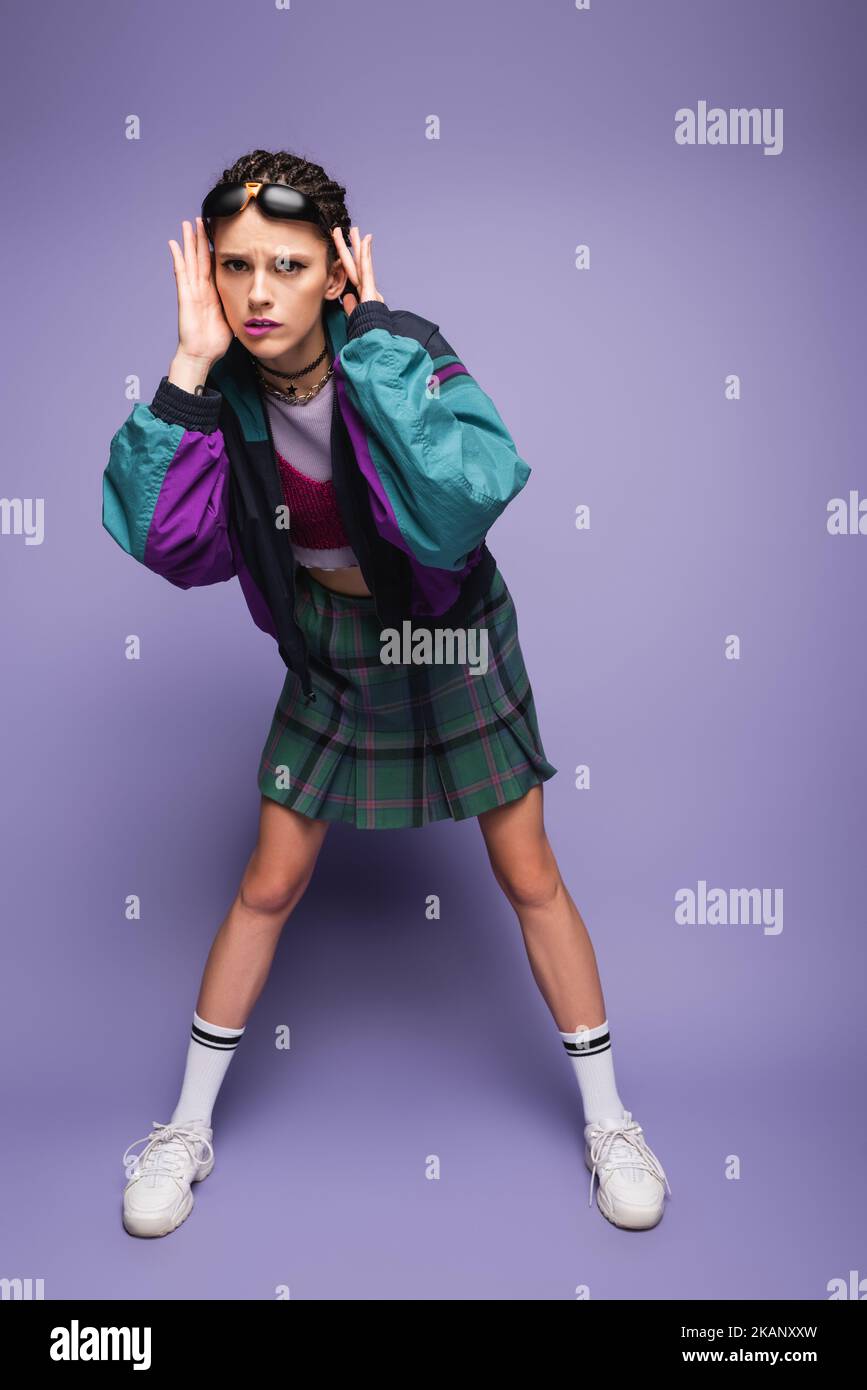 largo completo de mujer tensa en conjunto casual de los años noventa y sneakers blancas tocando la cabeza sobre fondo púrpura, imagen de stock Foto de stock