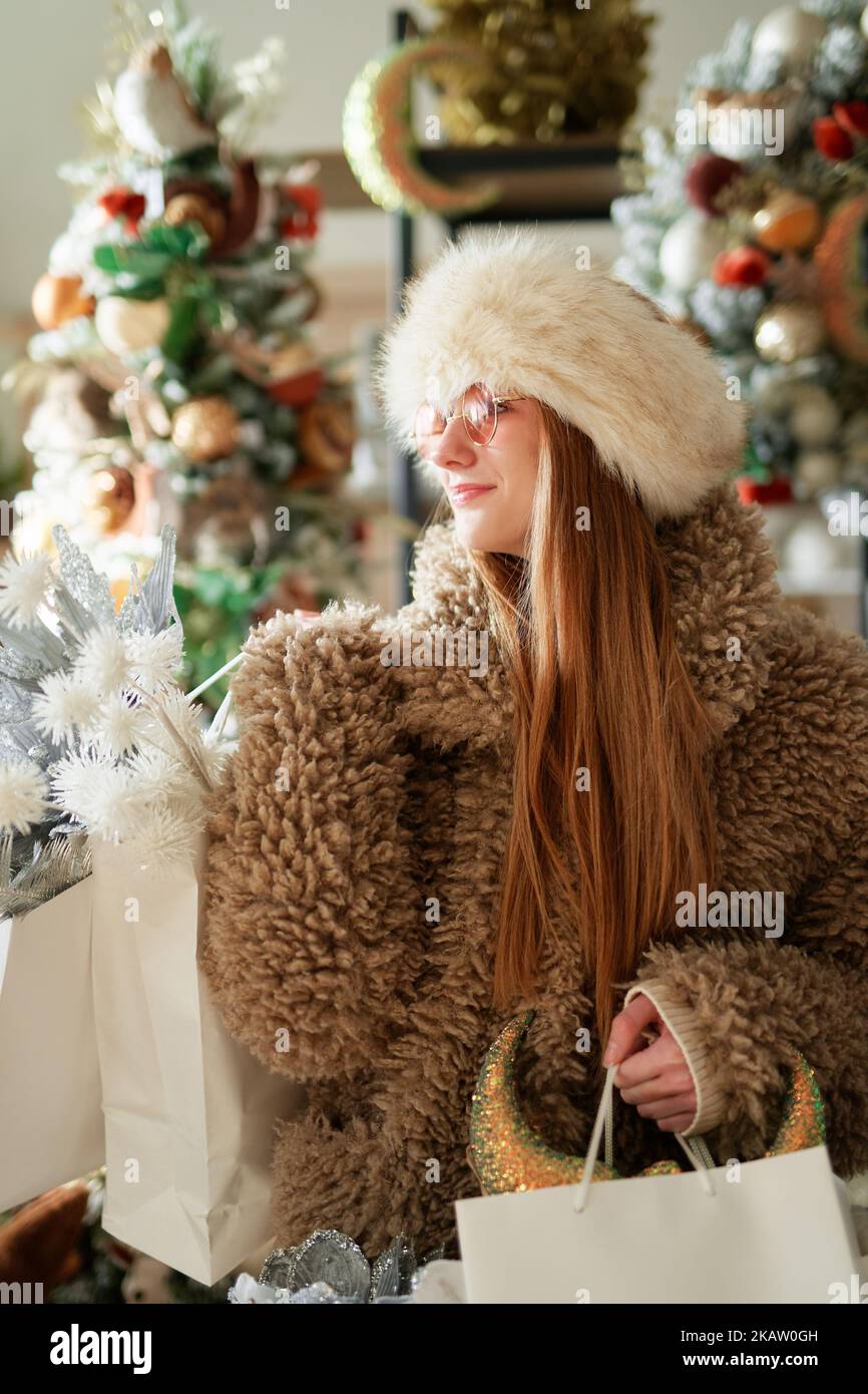 Adorable pelirroja caucásica en ropa de abrigo y gafas de sol con bolsas de compras llenas de decoración de Navidad en la tienda o supermercado con árbol de Navidad en el fondo. Tema de compras de Año Nuevo Foto de stock