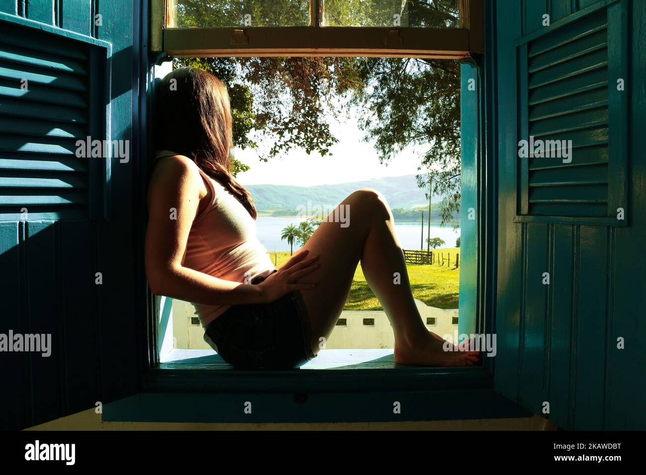 Mujer sentada en una ventana francesa mirando al exterior en un día soleado Foto de stock