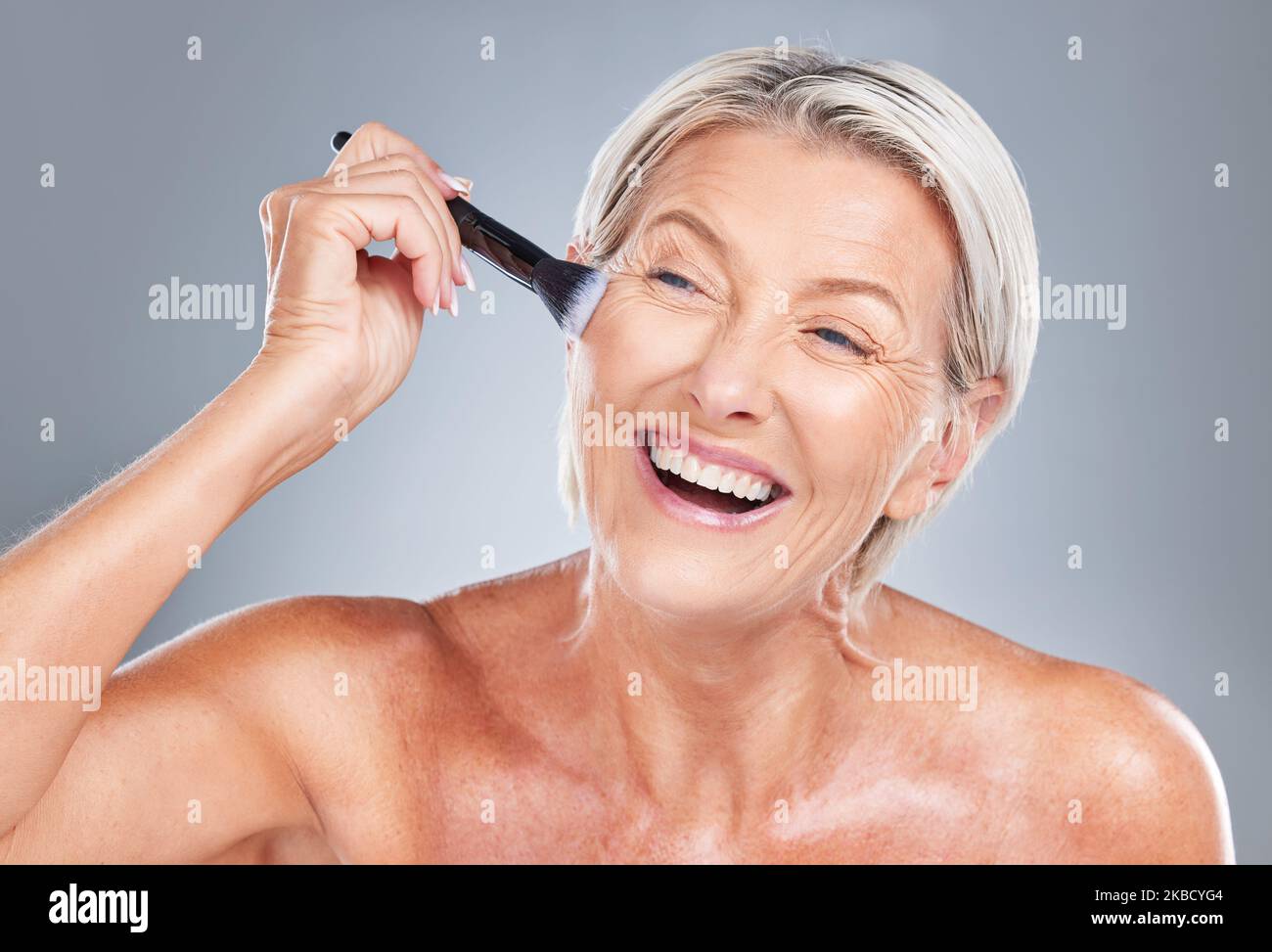Feliz, la mujer mayor y el cepillo de maquillaje en el estudio para la belleza, el cuidado de la piel y el tratamiento de las arrugas sobre un fondo gris. Cara, mejilla y producto de edad avanzada Foto de stock