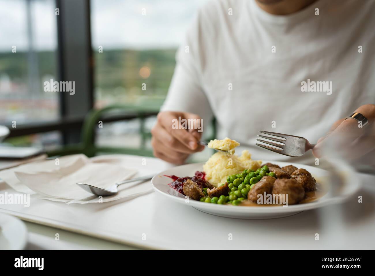 Primer plano en las manos de un hombre caucásico desconocido Siéntese a la mesa con tenedor y cuchillo Coma albóndigas con guisantes y puré de patatas Foto de stock