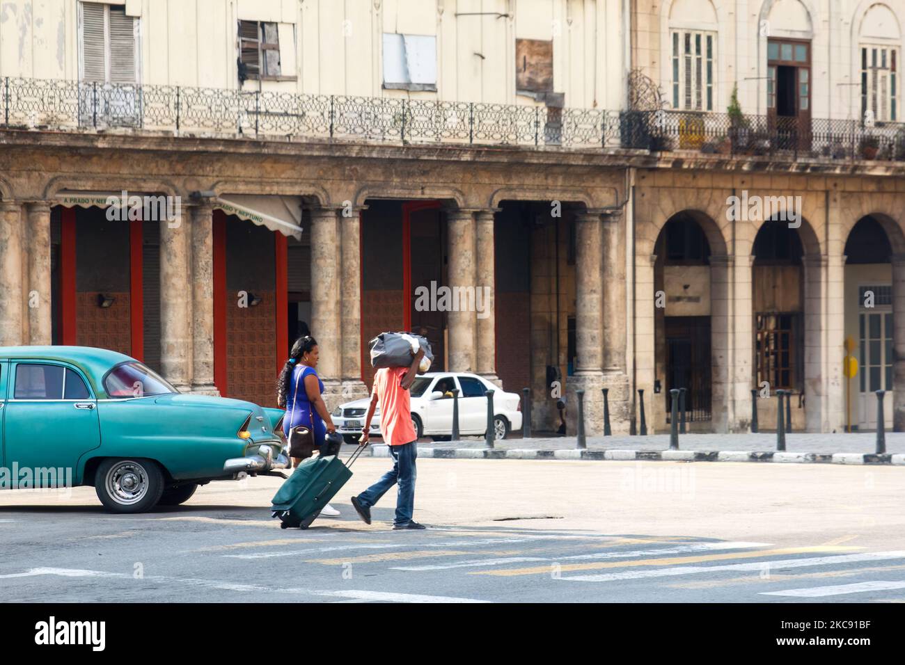 Los cubanos llevan dos grandes maletas en la calle de la ciudad. Un coche americano clásico conduce por fachadas de edificios erosionadas Foto de stock