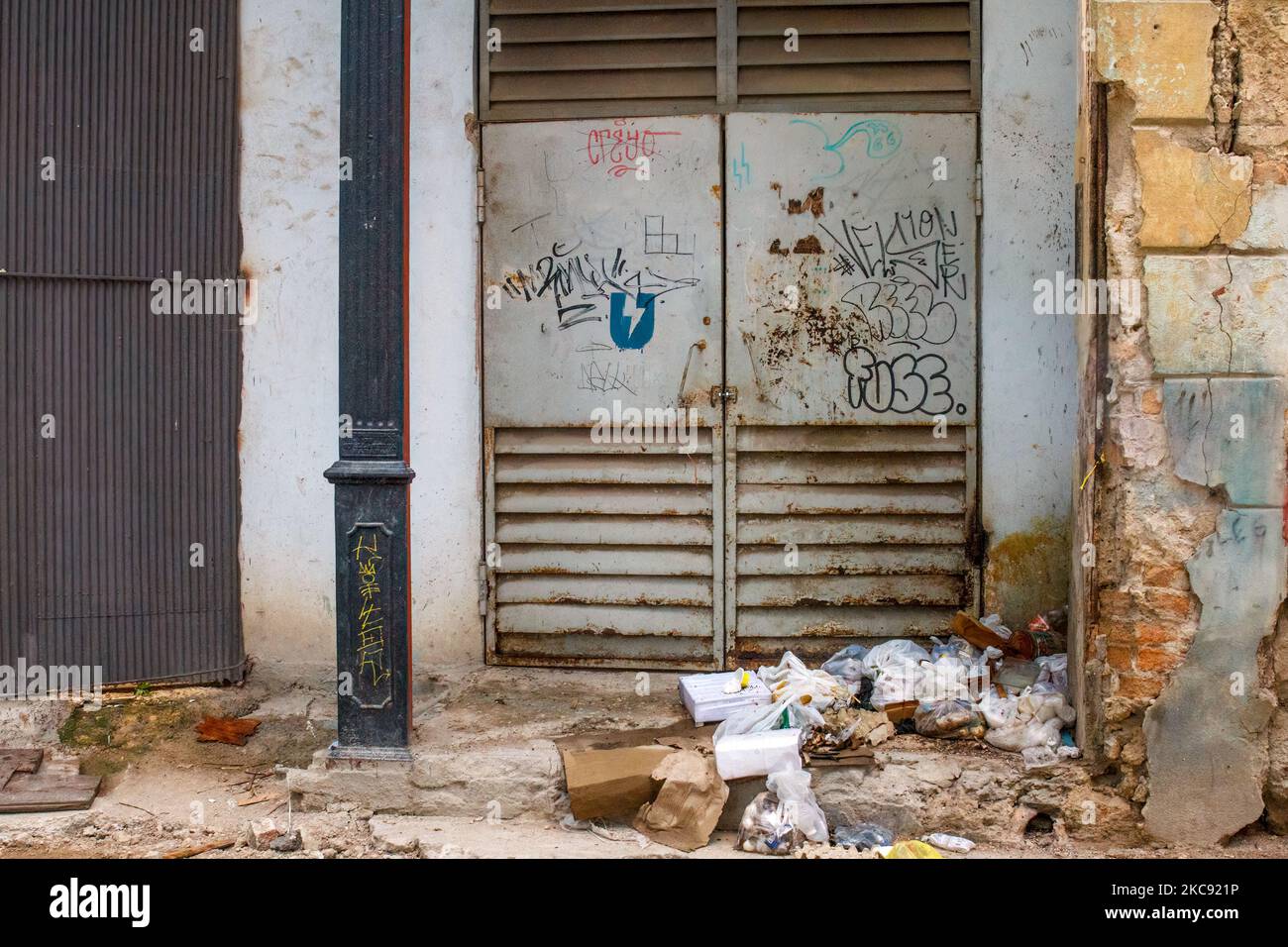Un montón de basura junto a una puerta metálica marcada con graffiti. Los edificios están ruinosos. Foto de stock