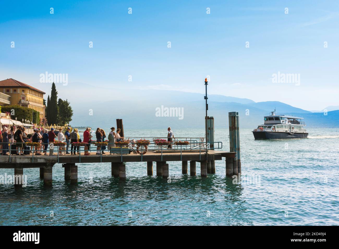 Lago Garda ferry, vista en verano de las personas de pie en el muelle de la estación de ferry en Gardone Riviera esperando la llegada de un ferry en barco, Lombardía, Italia Foto de stock