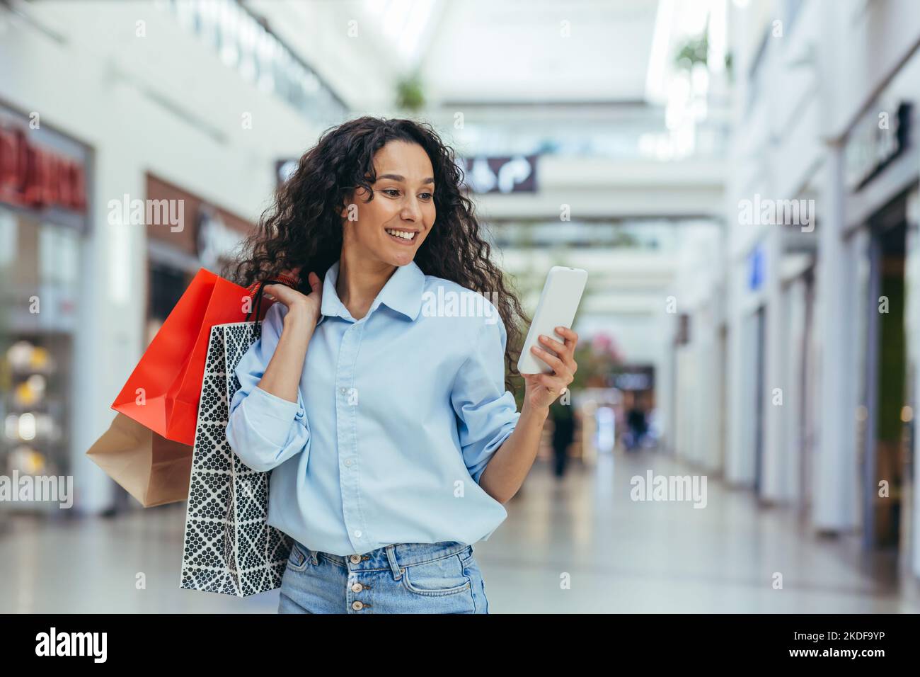 Mujer feliz comprando ropa durante el período de descuentos y ofertas promocionales, mujer hispana sonriendo y feliz navegando descuentos en línea utilizando el teléfono inteligente, dentro de una gran tienda moderna. Foto de stock