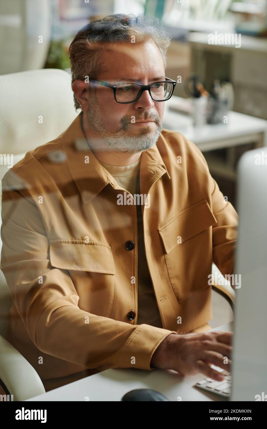 Moderno webdesigner maduro o programador sentado por escritorios frente al monitor de la computadora y mirando la pantalla mientras trabaja en línea Foto de stock