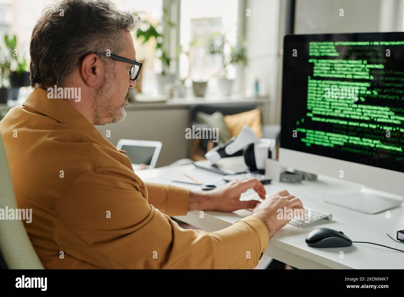Vista lateral de un ingeniero de TI serio y maduro que escribe en el teclado del ordenador mientras se sienta delante de la pantalla con datos codificados Foto de stock
