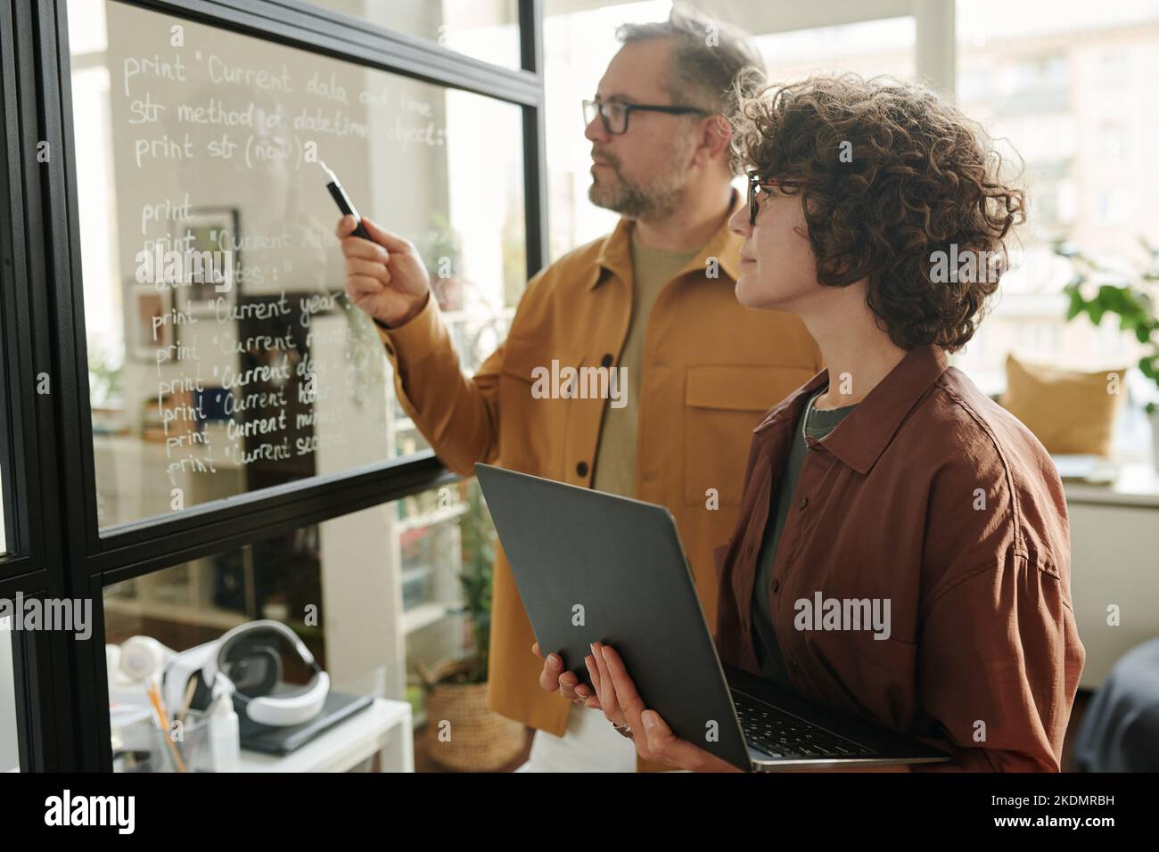 Mujer joven con un ordenador portátil mirando las notas en una pizarra transparente y escuchando a un compañero explicándolas durante la presentación Foto de stock