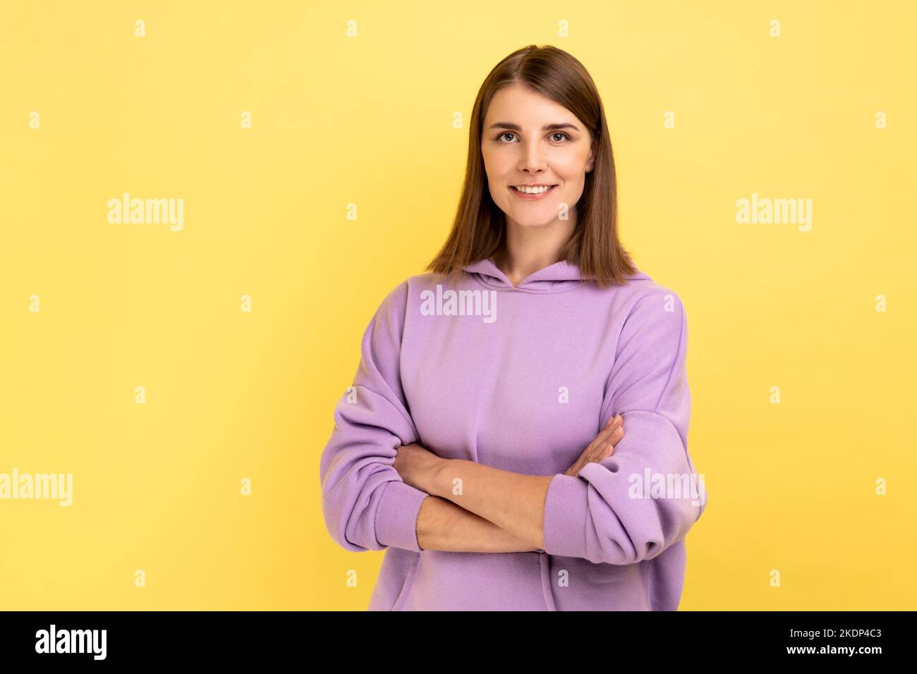 Retrato de feliz mujer exitosa de pie con las manos cruzadas, mirando la cámara con encantadora y alegre sonrisa tooty, usando sudadera con capucha púrpura. Estudio de interior grabado aislado sobre fondo amarillo. Foto de stock