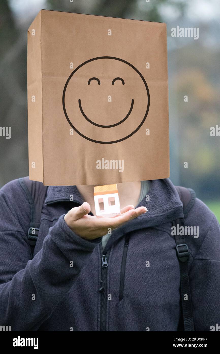 Hombre con una bolsa de papel sonriente en la cabeza. Un joven de pie sostiene un modelo de casa pequeña. La felicidad de comprar o alquilar una casa. Inmuebles. Foto de stock