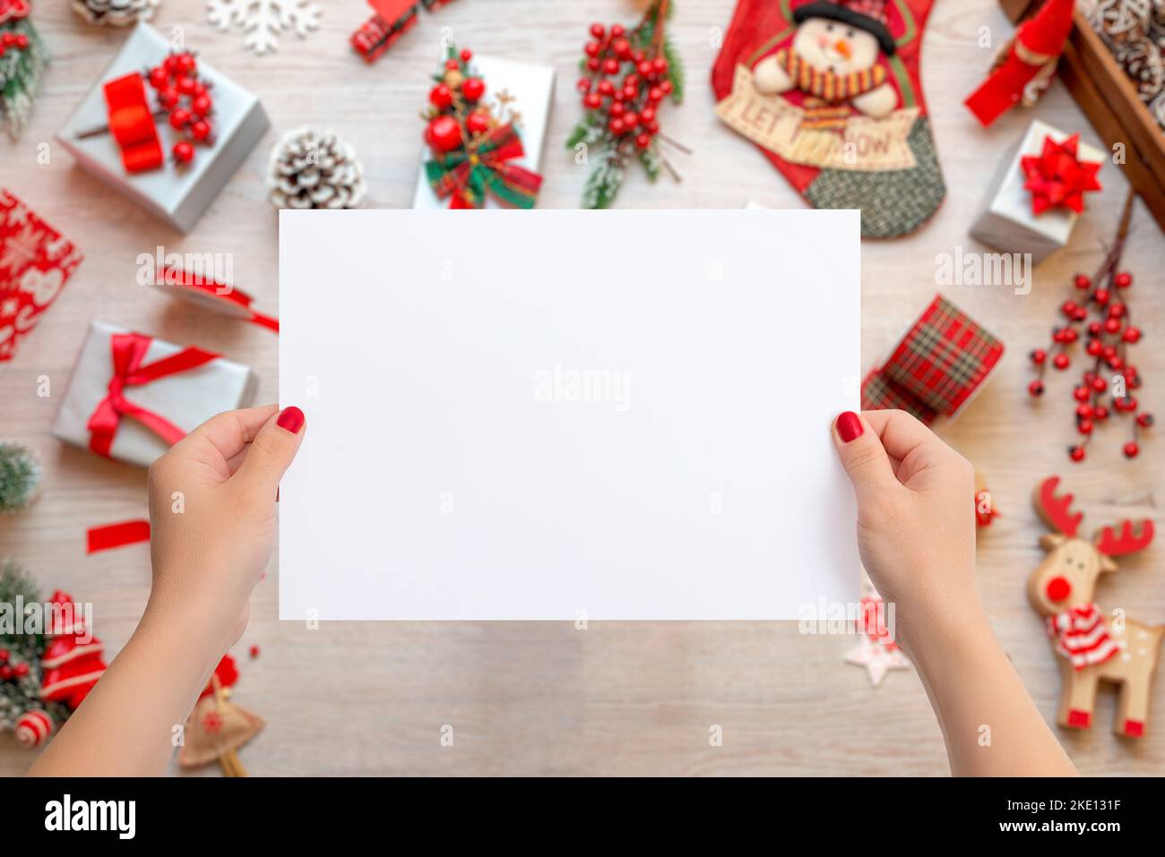 Mujer manos sosteniendo una hoja de papel en blanco para añadir texto de tarjeta de felicitación. Escena de Navidad con un montón de regalos y decoraciones sobre una mesa de madera Foto de stock