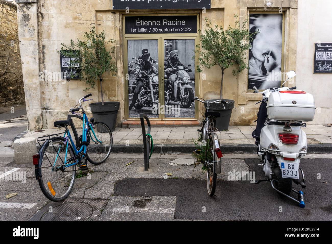 Dos bicicletas y una scooter aparcados delante de un cartel de una exposición de fotografía callejera de Etienne Racine en Arles, Francia. Foto de stock