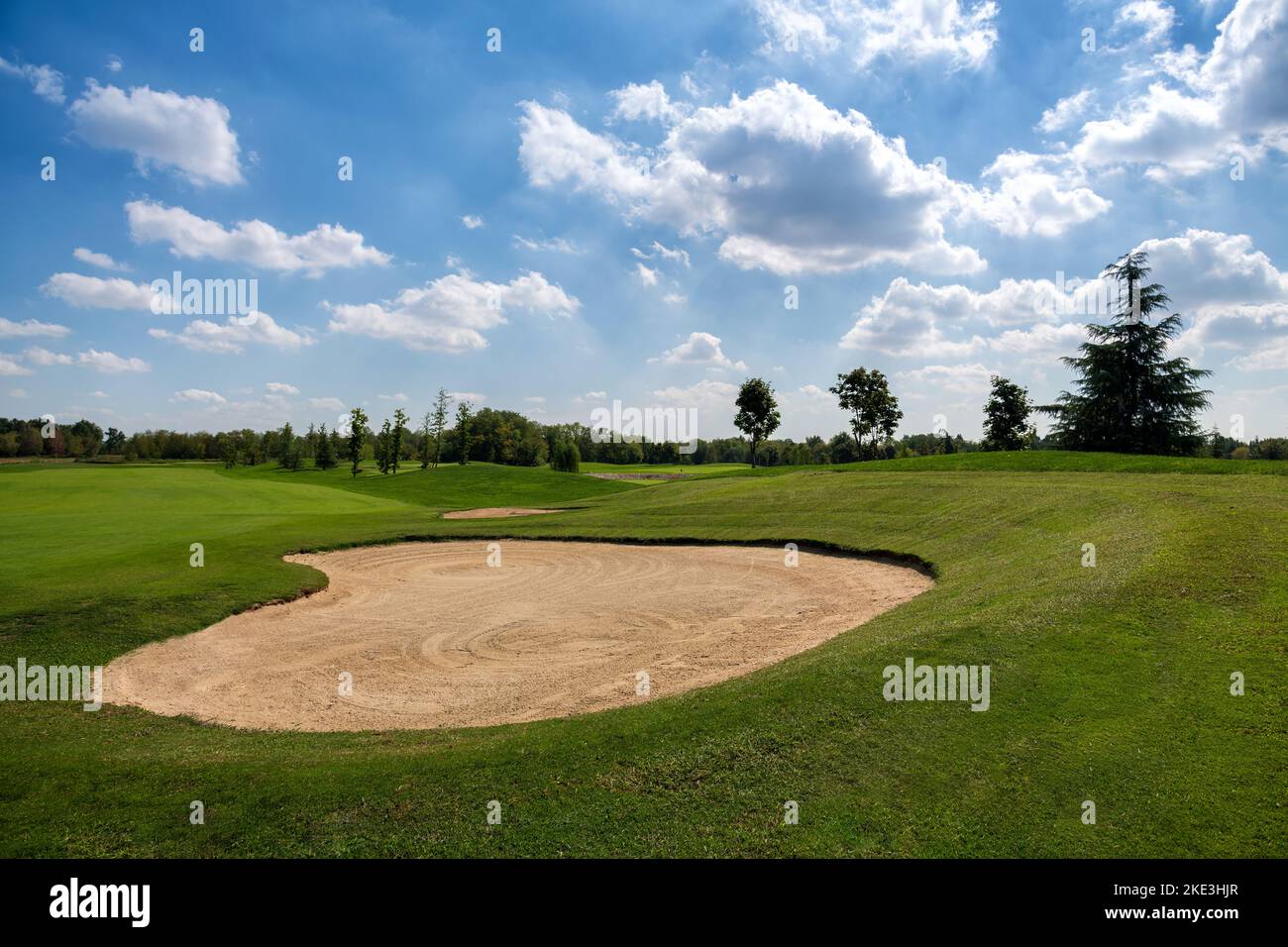 Trampa de arena rodeada de césped verde contra cielo azul nublado el día del fin de semana de verano en el campo de golf Foto de stock