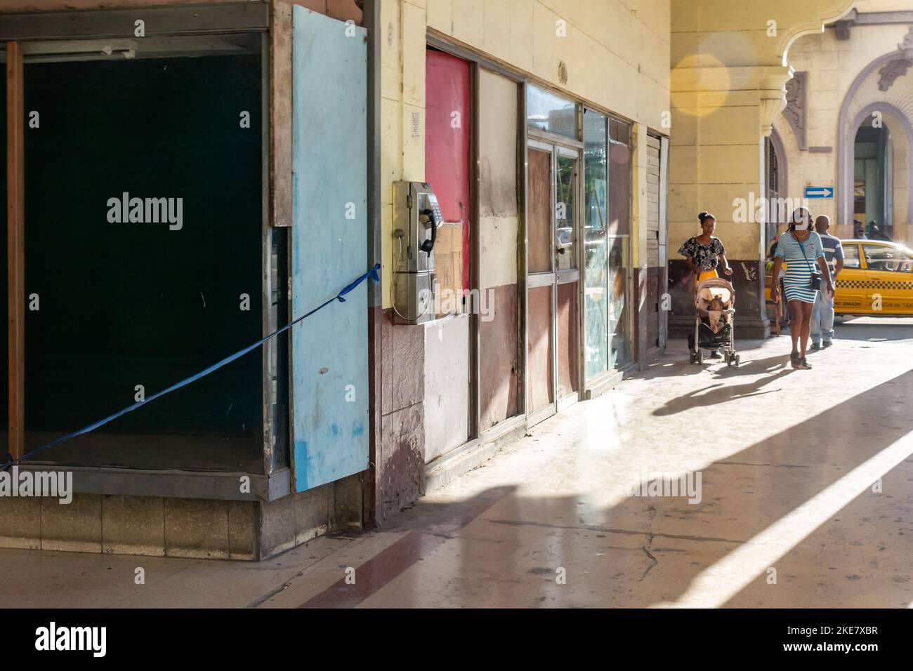 Los cubanos caminan debajo de un pórtico del edificio donde se proyectan sombras largas. Las puertas y ventanas se reparan con cartón. La pared exterior es dir Foto de stock