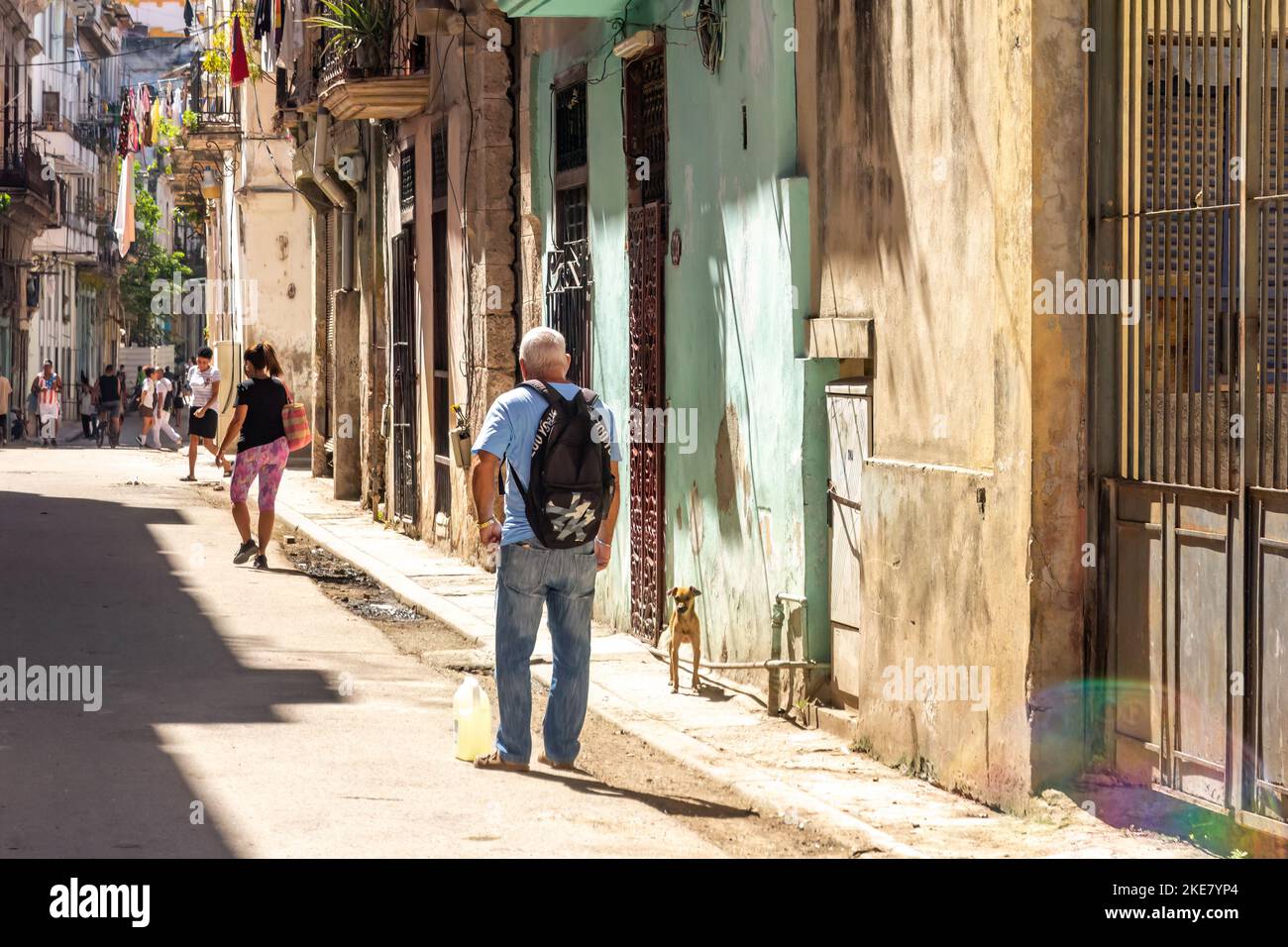 Un hombre cubano que lleva un contenedor de un galón lleno de líquido toma un descanso en un distrito residencial donde los edificios están desgastados y ruinosos. Foto de stock