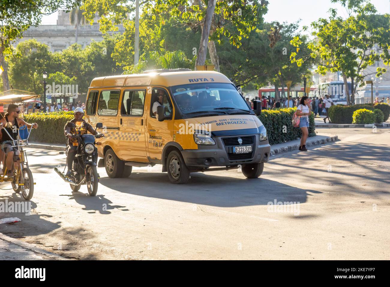 Una van Metrotaxi Lada conduce en una intersección de la ciudad donde una motocicleta y un bicitaxi también están en movimiento. Foto de stock