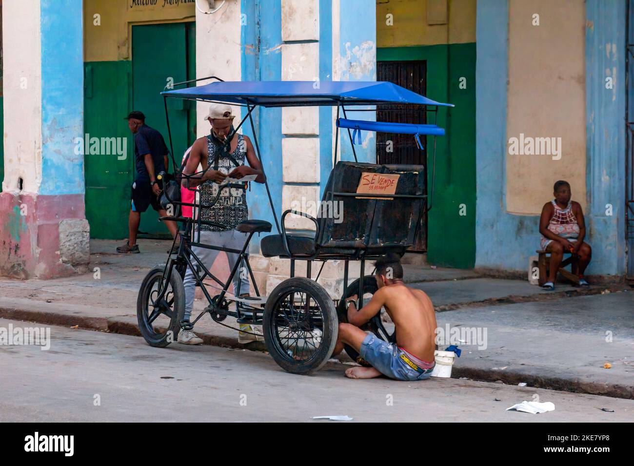 Un hombre cubano sin camisa reparando un bicitaxi o pedicab que tiene un letrero que dice 'En Venta' Foto de stock