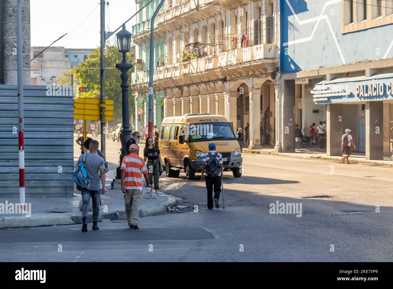 Los cubanos caminan en una intersección de la ciudad donde se ve una valla de construcción en la esquina. Un vehículo Lada con la inscripción Metrotaxi driv Foto de stock