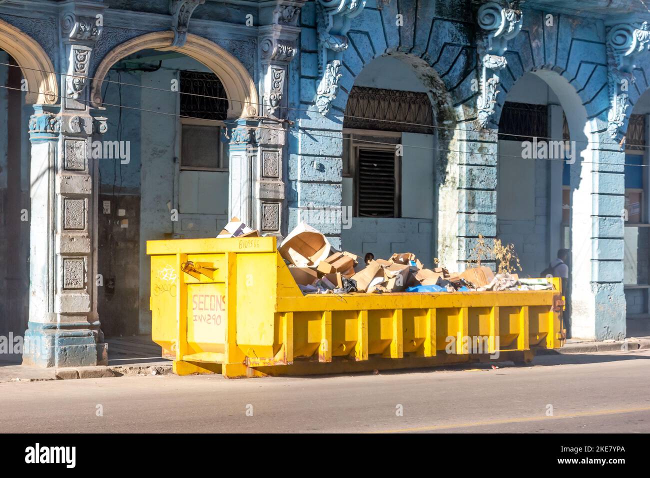 En la calle de una ciudad hay un contenedor metálico grande con el propósito de recoger basura. En el fondo hay un antiguo edificio arquitectónico. Foto de stock