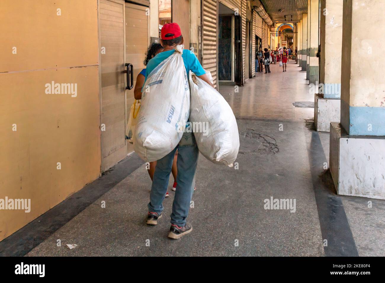 Un cubano lleva dos sacos pesados mientras camina en un porche de un edificio Foto de stock