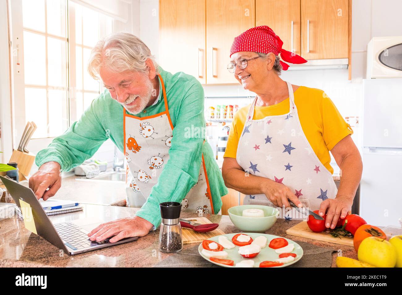 dos felices personas mayores cocinando juntos en la cocina aprendiendo y siguiendo un video tutorial de cocinar en el ordenador portátil - preparando juntos alimentos saludables Foto de stock